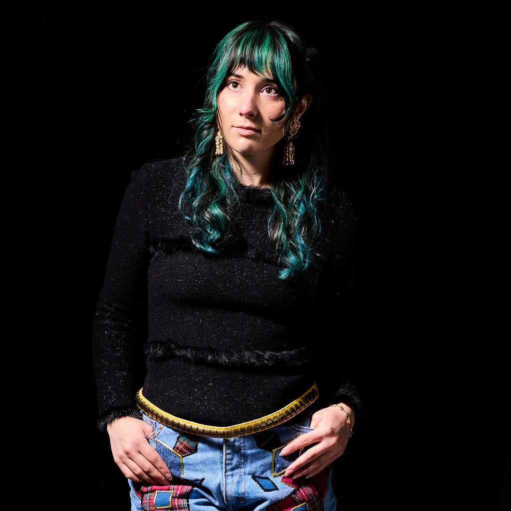 Person wearing a black top and colorful patterned pants standing against a dark background with decorative elements.