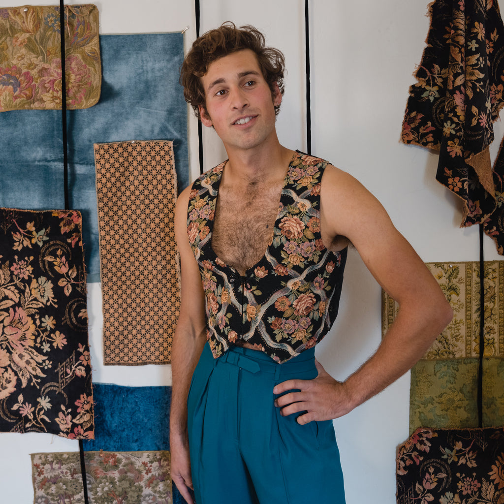 young man wearing a tapestry vest and blue high waisted pants, in front of a background of hanging fabric