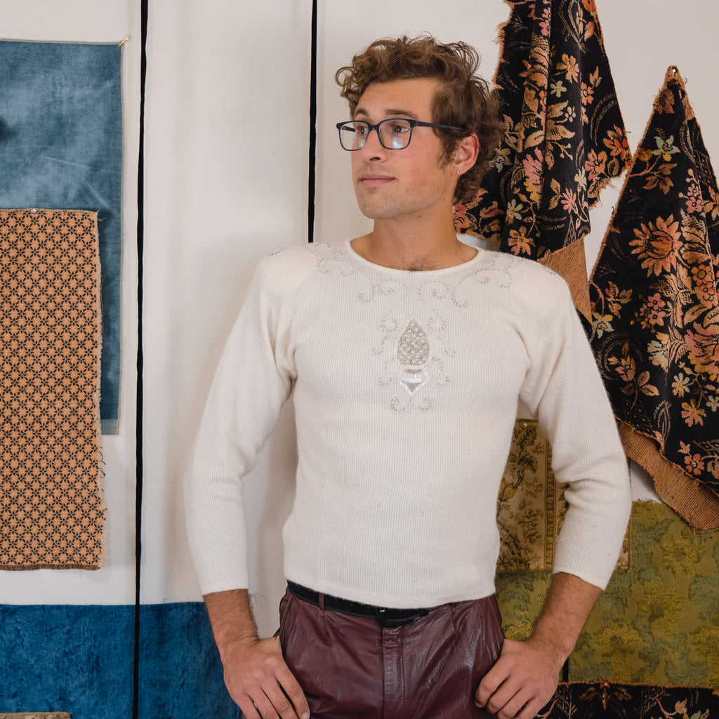 young man wearing a white beaded sweater with leather pants in front of a background with hanging fabric